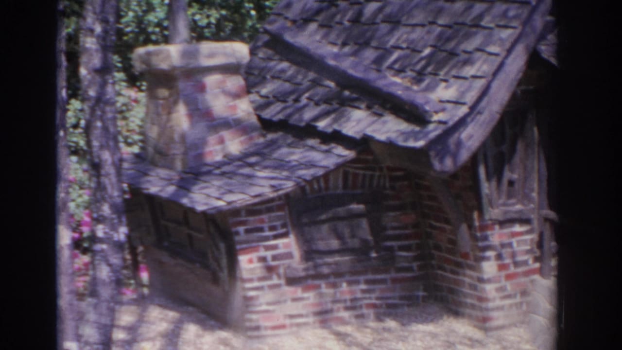 Miniature brick cottage with crooked roof, chimney, small window, and trees behind it.