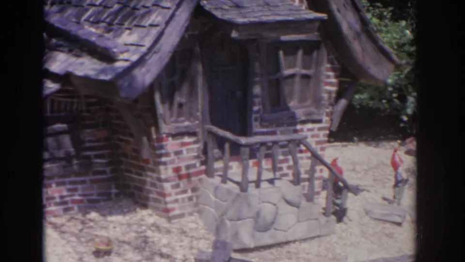 1962 Florida 8mm frame showing the miniature cottage entrance, porch rail, stone base, and tiny painted figures on the ground.