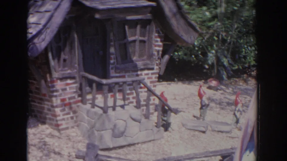 1962 Florida 8mm frame of a fairy tale-style miniature cottage side, with doorway, porch rail, stonework, trees, and tiny figures.