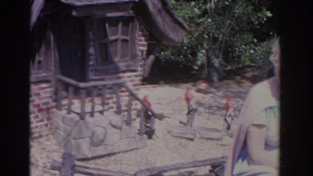 Woman seated near a stone bench beside a small gnome house and garden figures.
