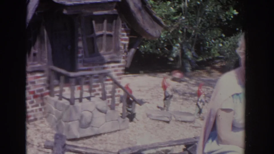 1962 Florida 8mm frame showing a seated woman near a stone bench, novelty gnome house, and garden figures in bright sun.