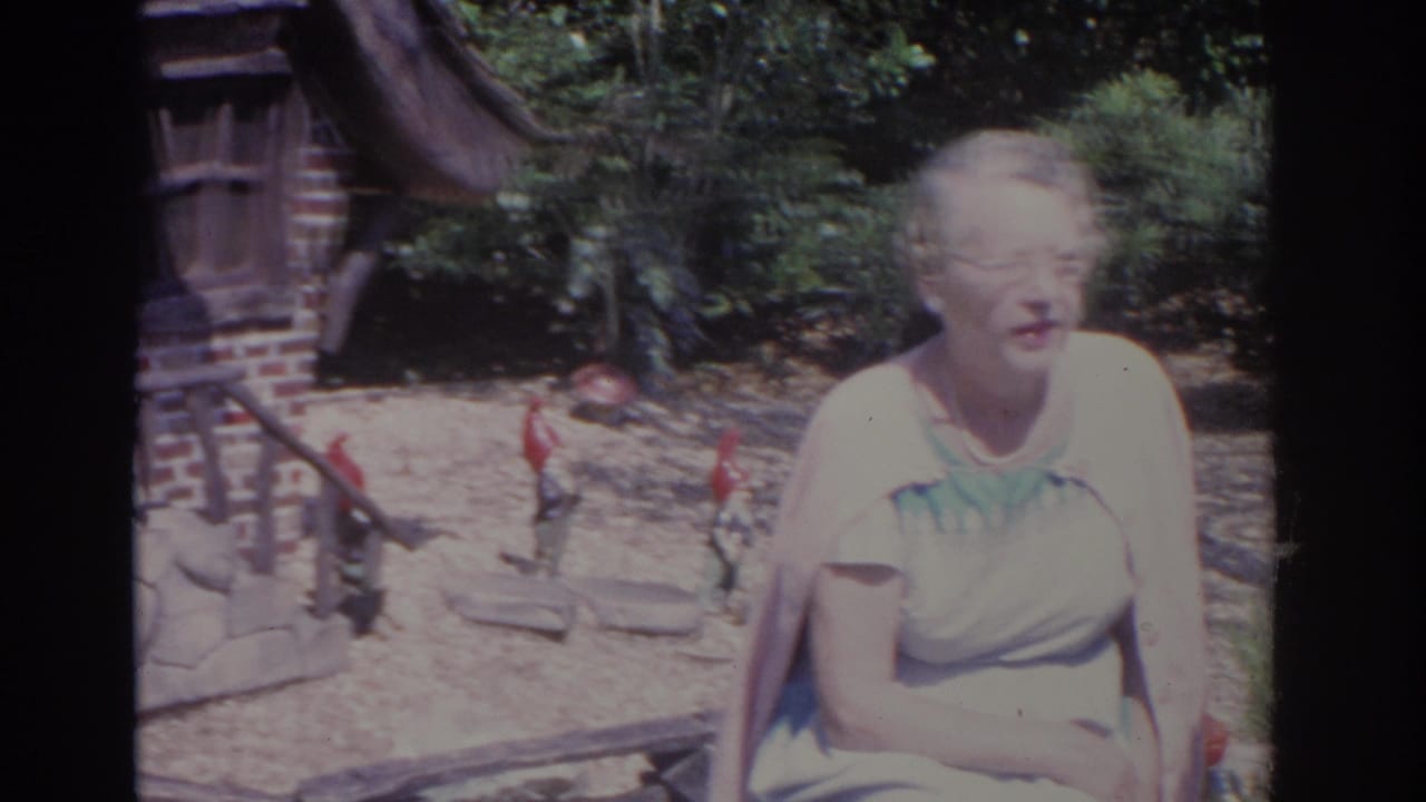 Woman in a pale blue dress sits in front of shrubs and a gnome house.
