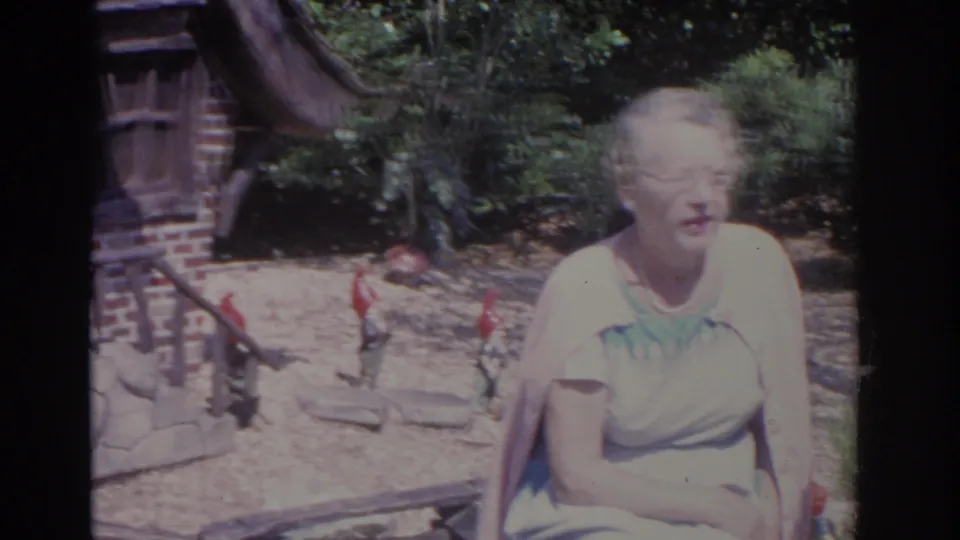 1962 Florida home-movie frame with a woman in a pale blue dress seated near shrubs, gnomes, and a small brick-and-wood house.
