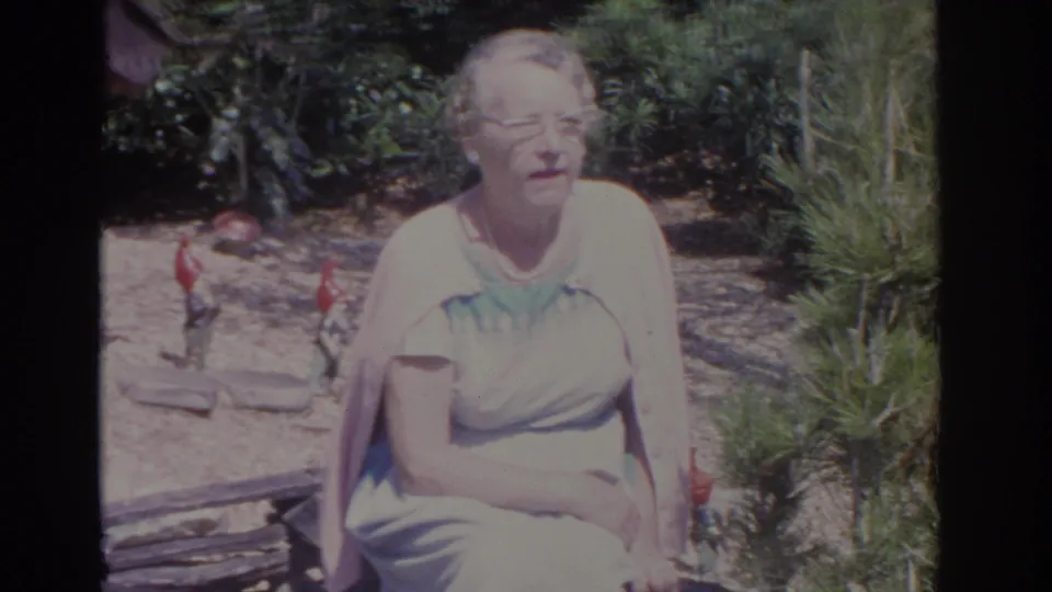 1962 Florida 8mm frame showing a seated woman on a stone bench, with garden gnomes, shrubs, and sunlit landscaping behind her.