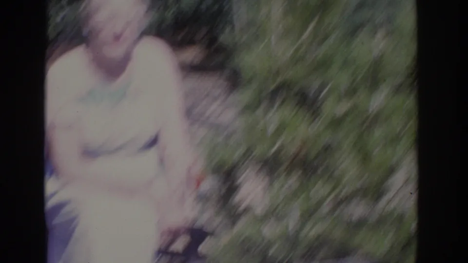 Soft, blurred 1962 Florida 8mm frame of a seated older woman in a pale dress, partly obscured by camera movement and foliage.
