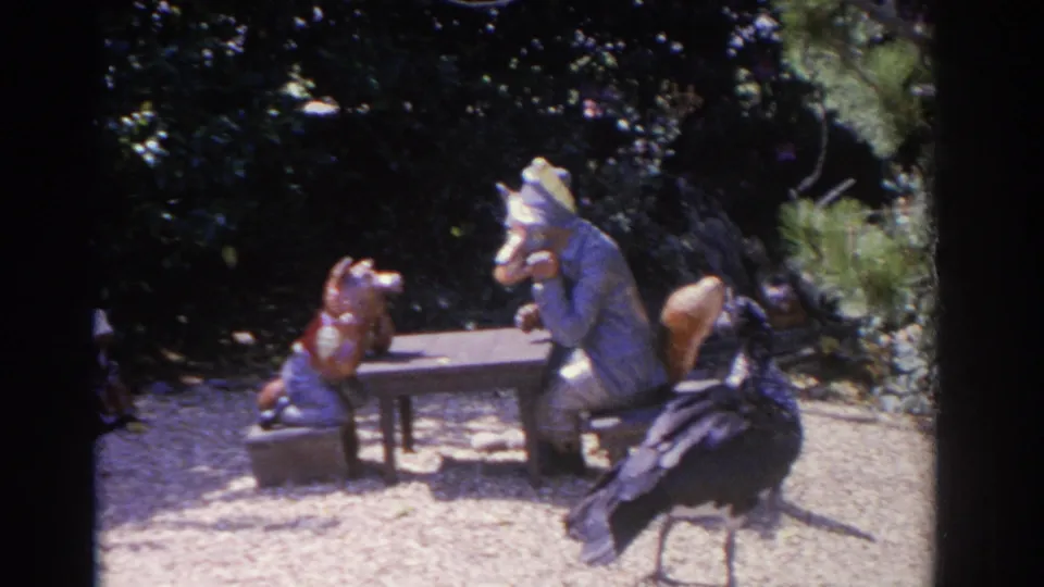 1962 Florida 8mm frame showing masked costumed figures around a small table as a large dark bird stands in the sunlit garden.