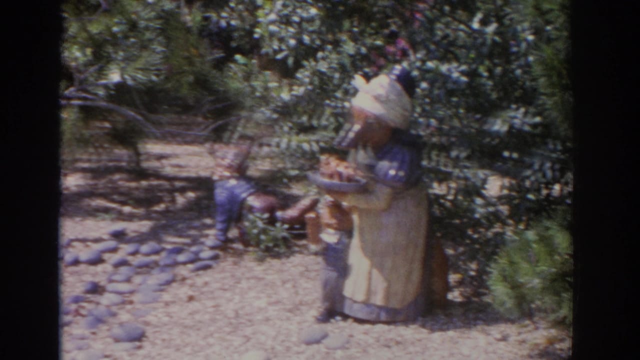 Carved bear-like statue in an apron beside foliage and a smaller wooden figure.