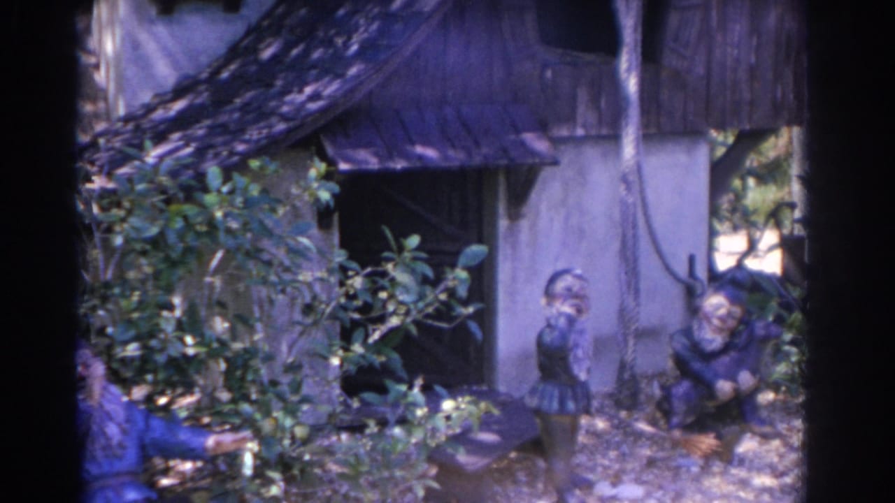 Garden gnomes arranged beside a small cottage with shrubs in the foreground.
