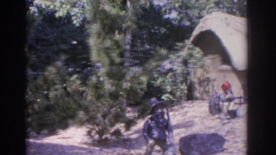 1962 Florida 8mm frame showing a gnome figure, thatched cottage, small fence, shrubs, and sandy ground in a whimsical garden village display.