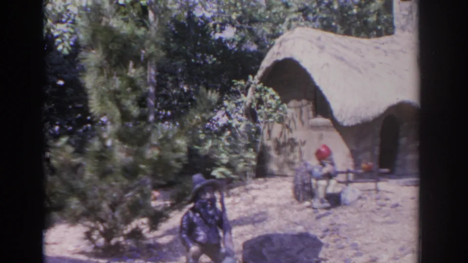 1962 Florida 8mm frame with gnome figures near a small thatched cottage, shrubs, fence pieces, and sandy ground in a decorative outdoor display.