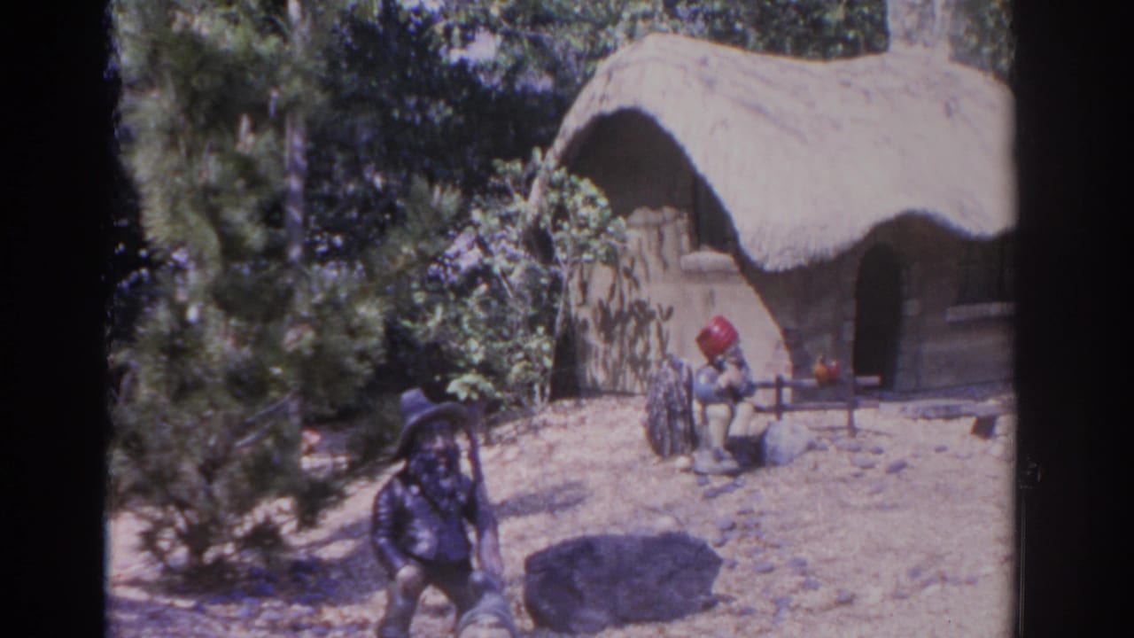 Two gnome figures stand near a small thatched hut in a sandy garden setting.