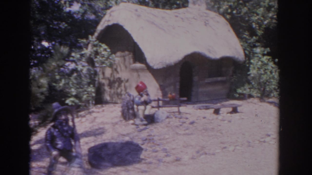 Gnome figures and a thatched hut appear in a sunlit sandy yard.
