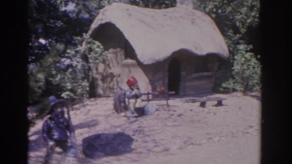 1962 Florida home-movie frame with garden gnomes near a rustic thatched hut, bordered by rocks, low fencing, and leafy vegetation.