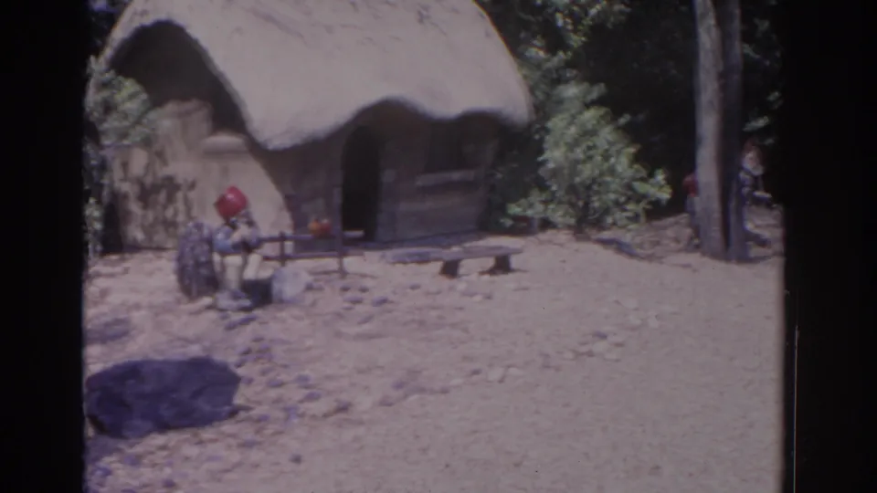 1962 Florida 8mm frame showing a red-hatted garden gnome by a thatched hut, with a second figure partly cropped and trees in the background.