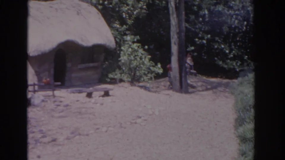 1962 Florida home-movie frame centered on the thatched hut, with a partly cropped gnome, bench, trees, and sandy ground visible.