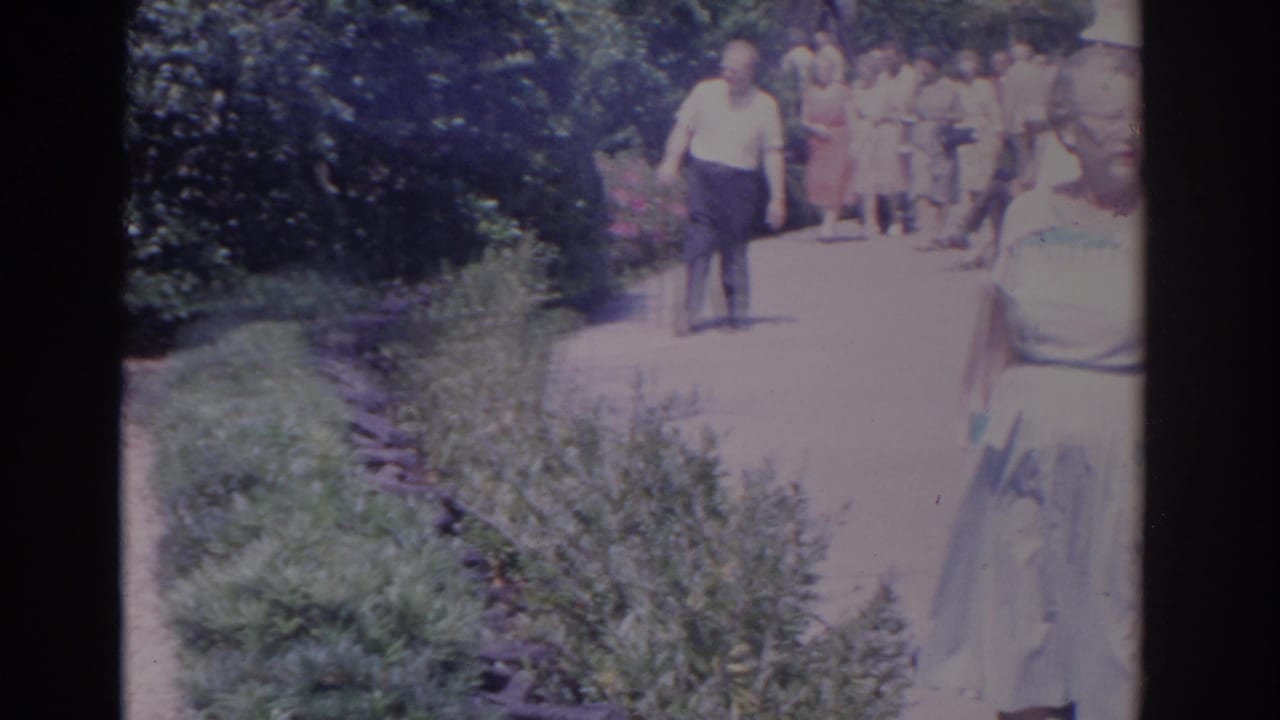 Adults in 1960s attire walk along a bright garden path bordered by greenery.