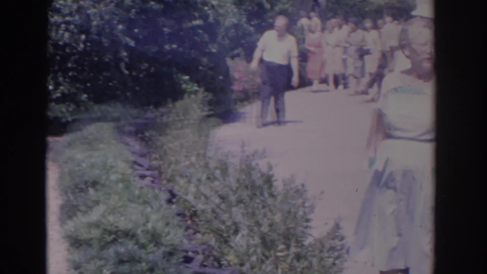 1962 Florida home-movie frame of adults in suits and dresses walking along a sunlit garden path.