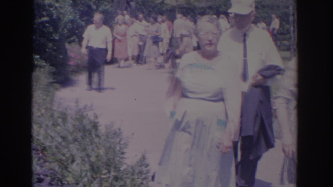 1962 Florida home-movie frame with adult visitors passing the camera on a garden walkway.