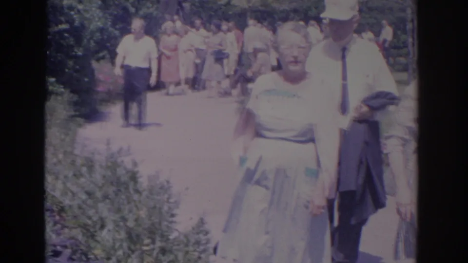 1962 Florida home-movie frame with adult visitors passing the camera on a garden walkway.