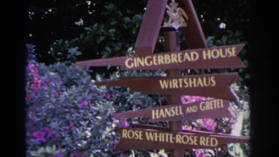 1962 Florida frame of a wooden fairy-tale attraction sign partly surrounded by flowers and foliage.