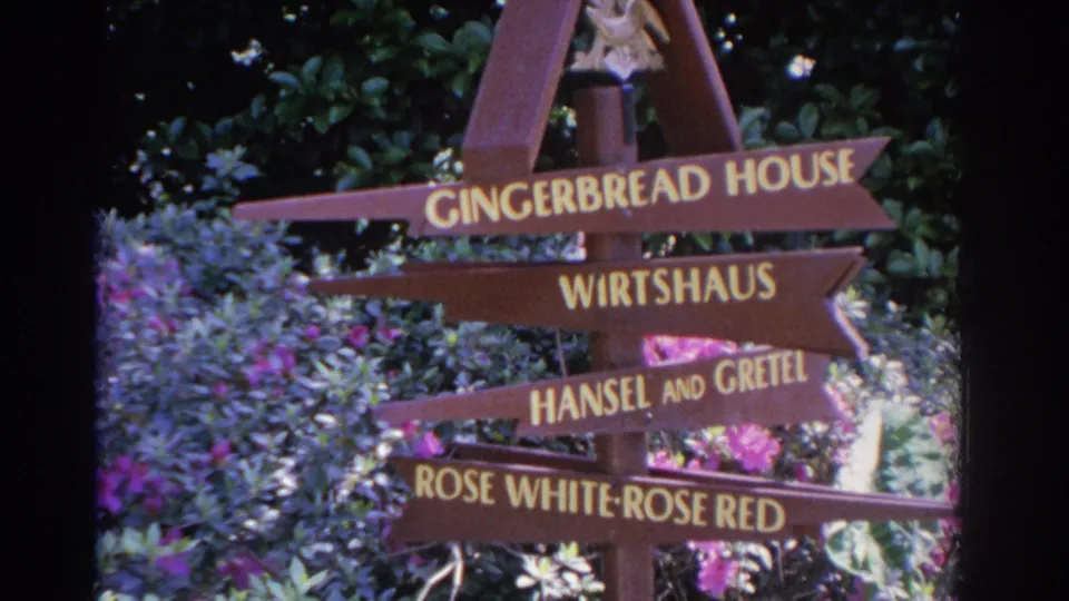 1962 Florida home-movie frame showing a readable fairy-tale themed directional sign in a garden setting.