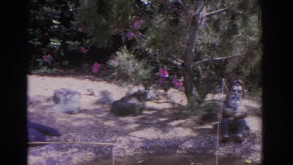 1962 Florida 8mm frame showing a landscaped garden pond with rocks, shrubs, pink flowers, and a small seated gnome figure.