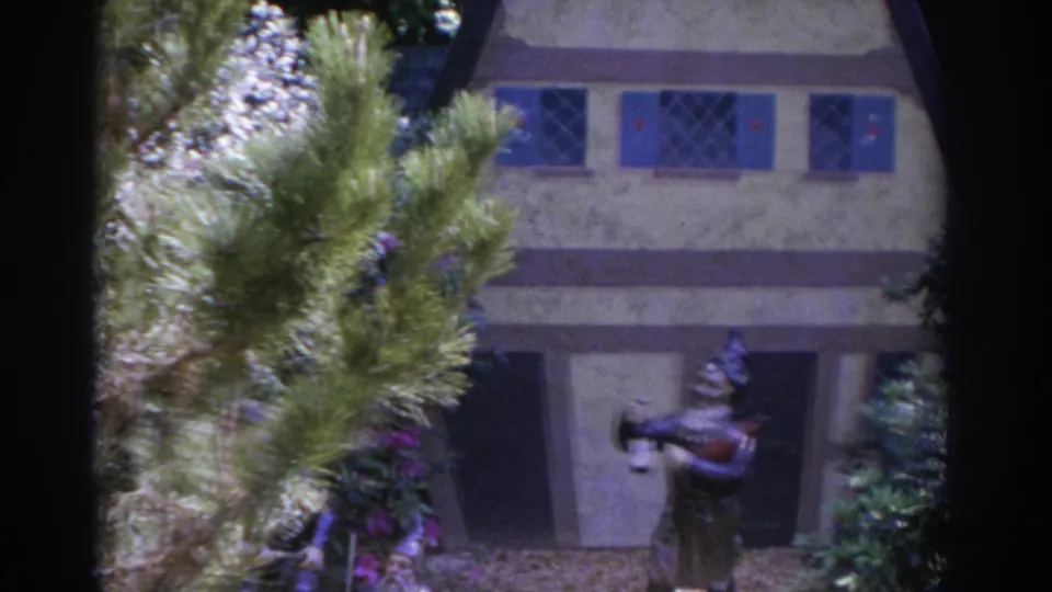 1962 Florida 8mm frame of a miniature house facade with blue shutters, plants in front, and a garden figure near the doorway.