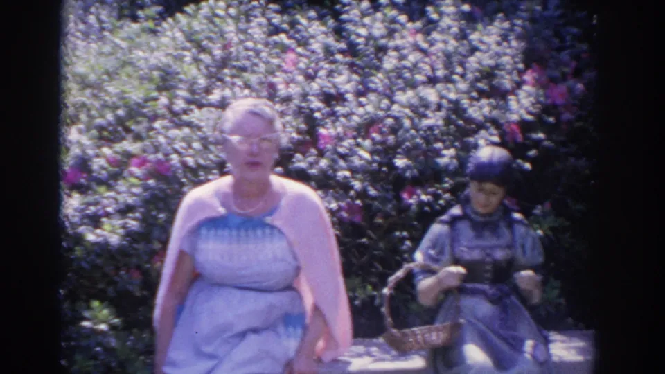 1962 Florida home-movie frame showing an older woman seated outdoors beside a dark ornamental garden statue and flowering shrubs.