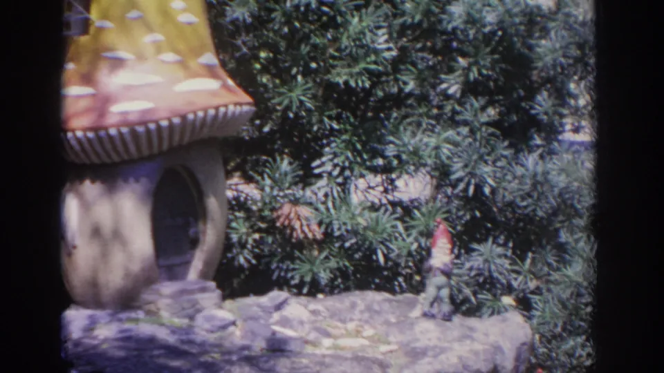 1962 Florida home-movie frame of the mushroom-shaped garden house with pine foliage and a small gnome figure nearby.