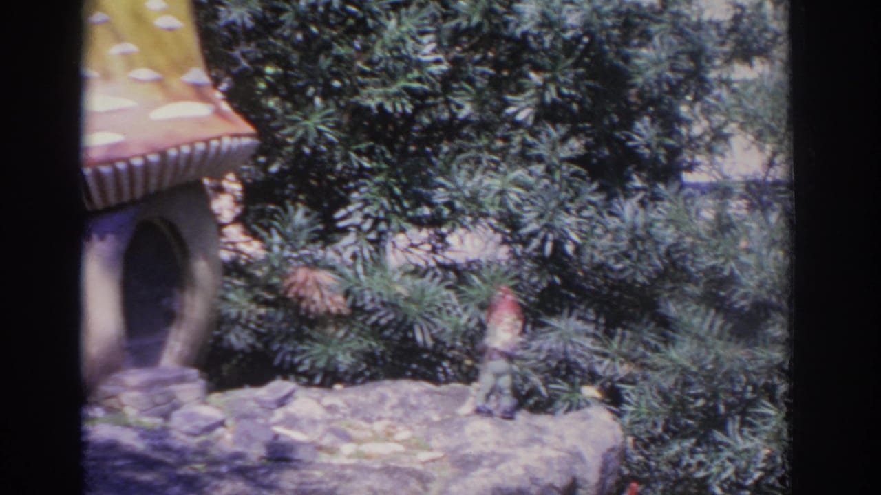 Garden gnome beside a small mushroom-shaped house among shrubs and rocks.