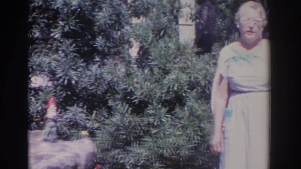 1962 Florida 8mm frame showing an older woman in a light dress at the edge of a garden with a gnome figure nearby.
