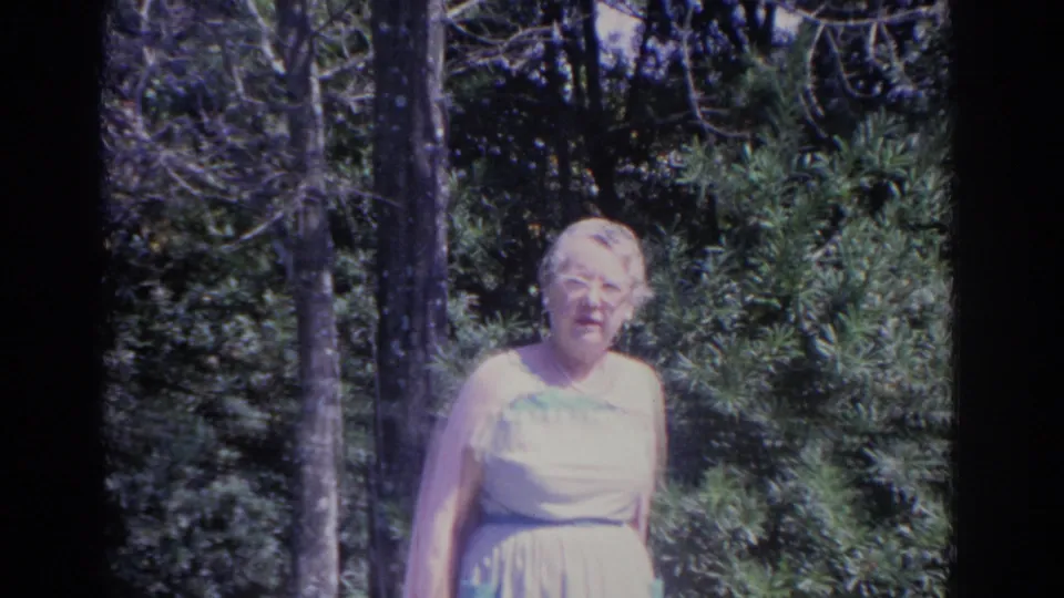 1962 Florida home-movie frame of an older woman standing in a shaded garden area with trees and shrubs behind her.