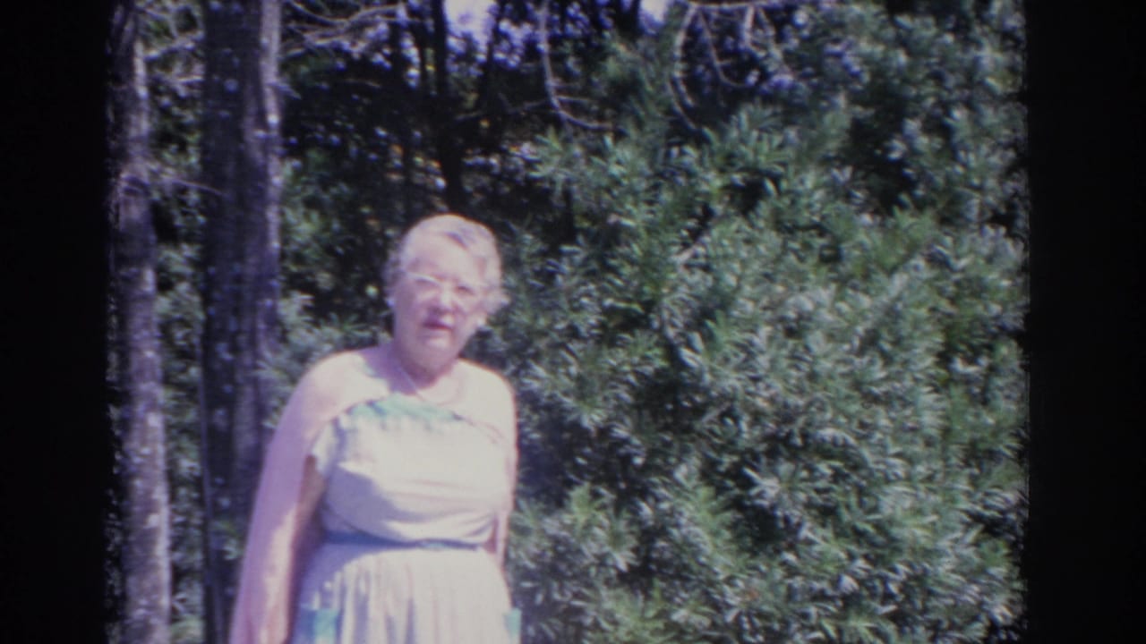 Elderly woman in glasses stands beside dense garden shrubs and trees.