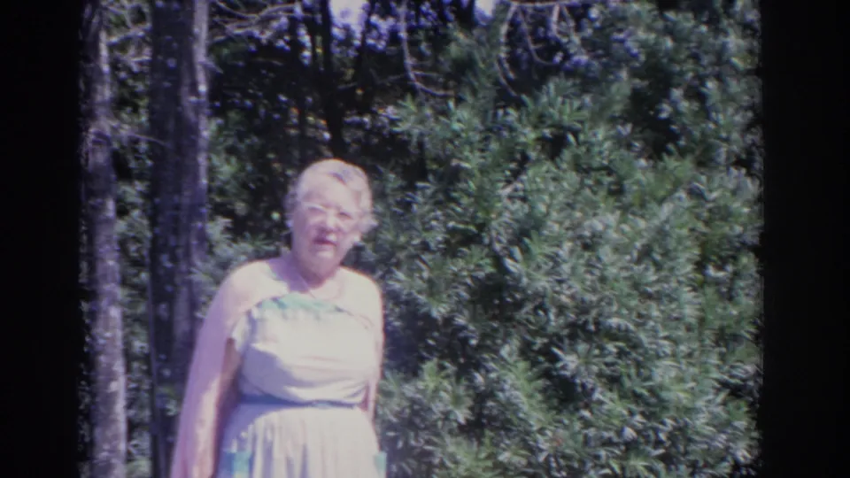 Elderly woman in a light dress and glasses stands in a lush Florida garden in 1962 home-movie footage.