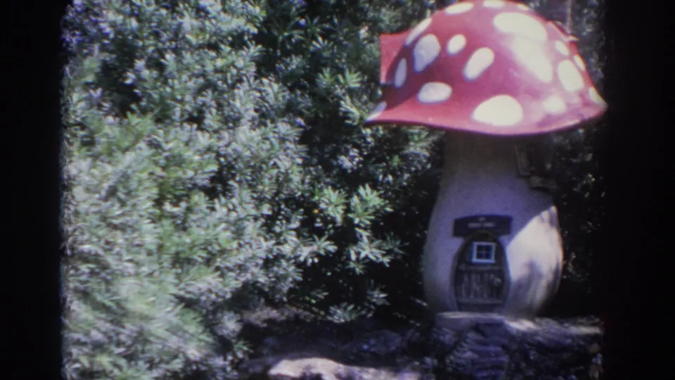 A whimsical mushroom-shaped garden house with a red spotted cap and small dark door stands among dense greenery.