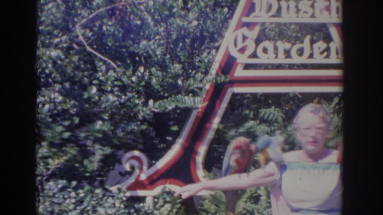 Woman with glasses holds parrots near a large Botanical Gardens sign and leafy background.