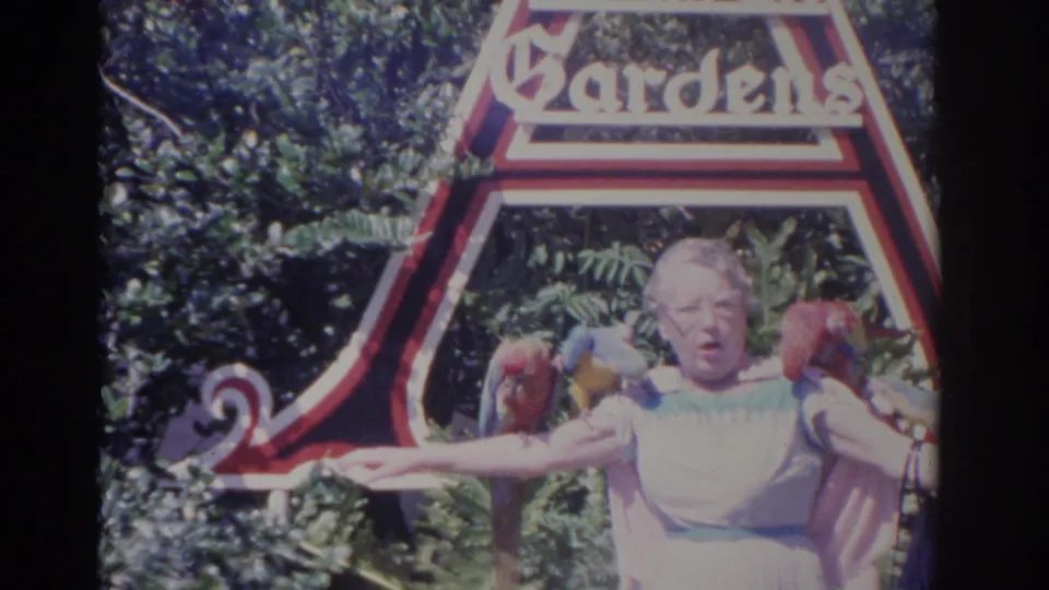 1962 Florida home-movie still showing an elderly woman posing with parrots on her arms and shoulder beneath the Botanical Gardens sign.