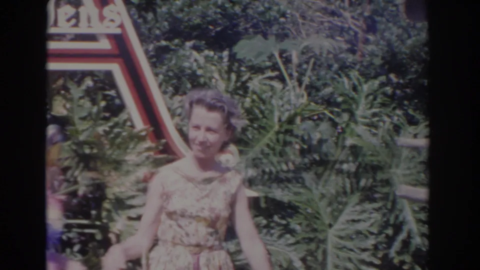 A woman in a floral dress smiles near the partially visible Busch Gardens sign, with tropical greenery and a macaw at the frame edge.
