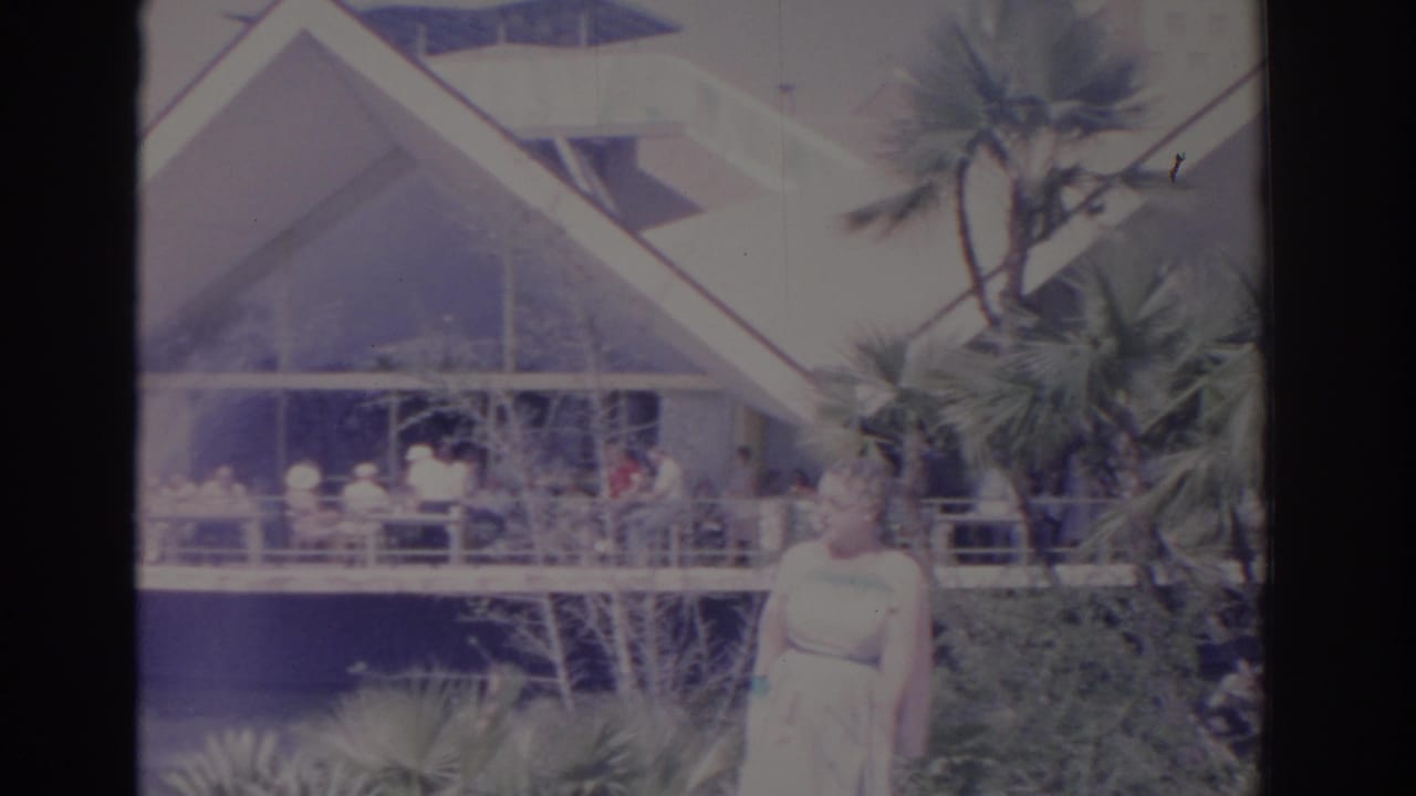 Woman in a light dress stands before palms and an angular building.
