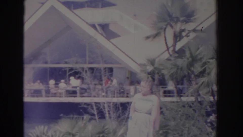 1962 Florida home-movie frame showing a woman near palm landscaping and a modernist-looking building.