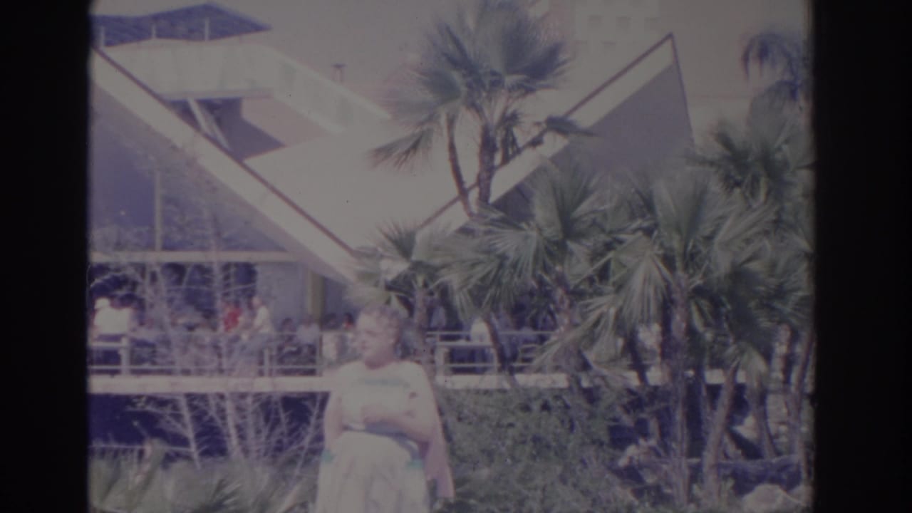 Woman stands near palms with a bright angular building behind her.