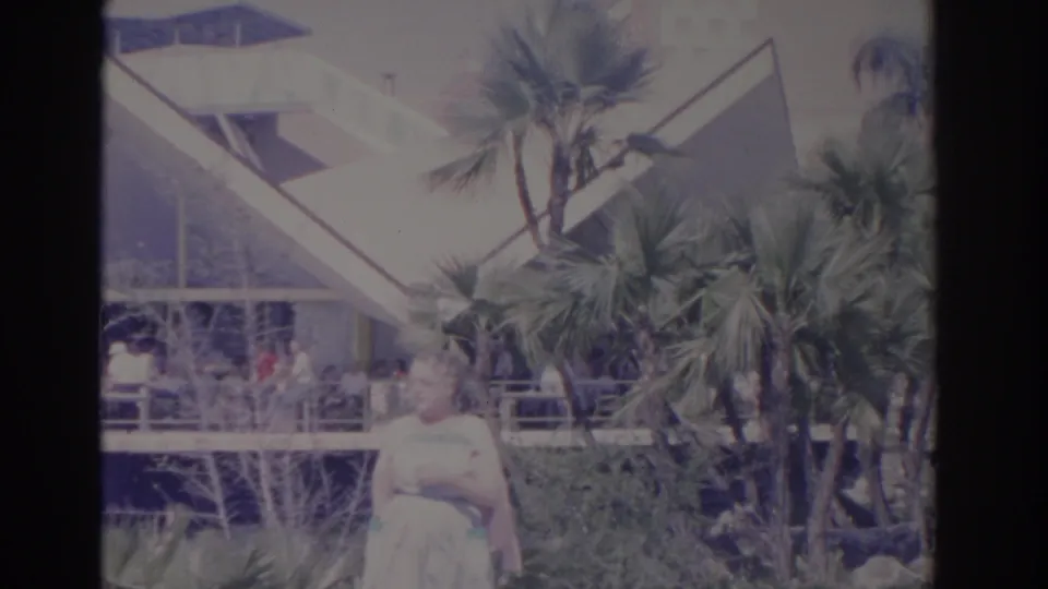 1962 Florida footage frame with a woman in the foreground, palm trees, railings, and a bright angular building behind.