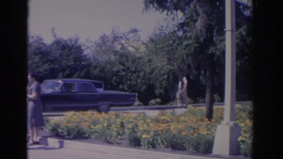 1962 Florida 8mm frame showing a dark sedan by yellow-orange flower beds, with a woman standing at the left curb.