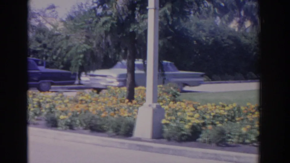 1962 Florida 8mm frame with two period cars near roadside flowers and a man walking along the sidewalk.