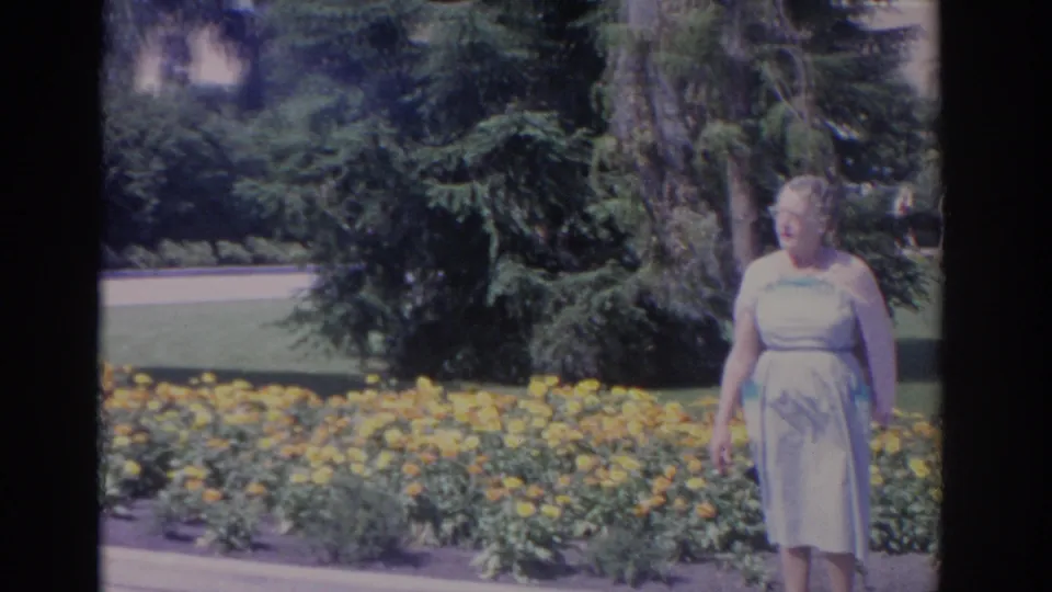 An adult woman in a pale dress walks near yellow flowers, lawn, trees, and a roadside garden in 1962 Florida home-movie footage.