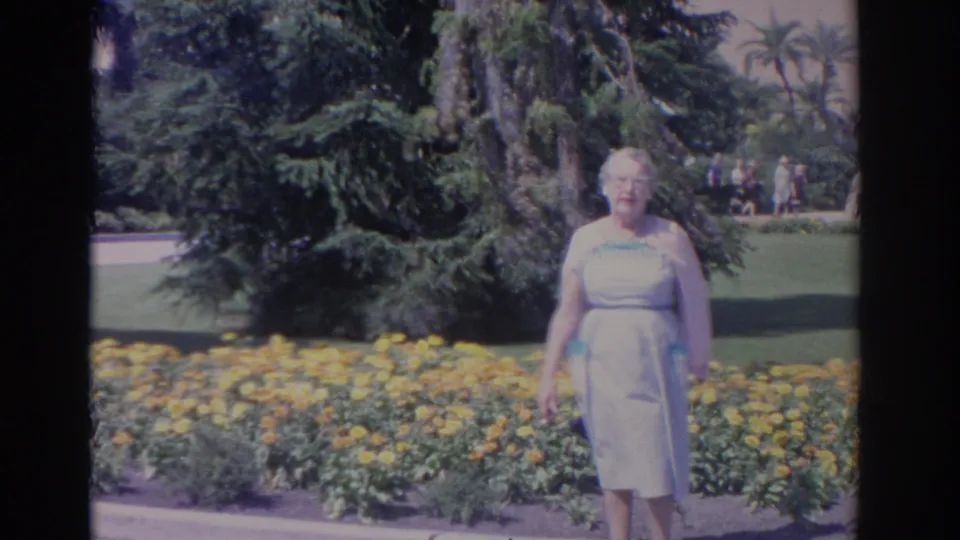 In 1962 Florida home-movie footage, a woman in a pale blue dress stands near a yellow flower bed with lawn, trees, and palms behind her.
