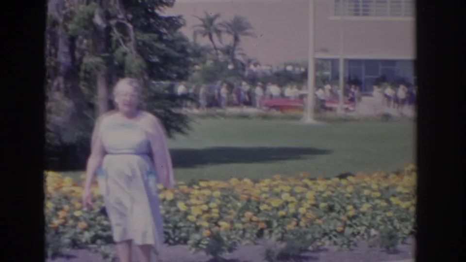 The handheld frame shifts left as the woman stands near the flower bed; a building, distant visitors, palms, and a red car appear behind her.