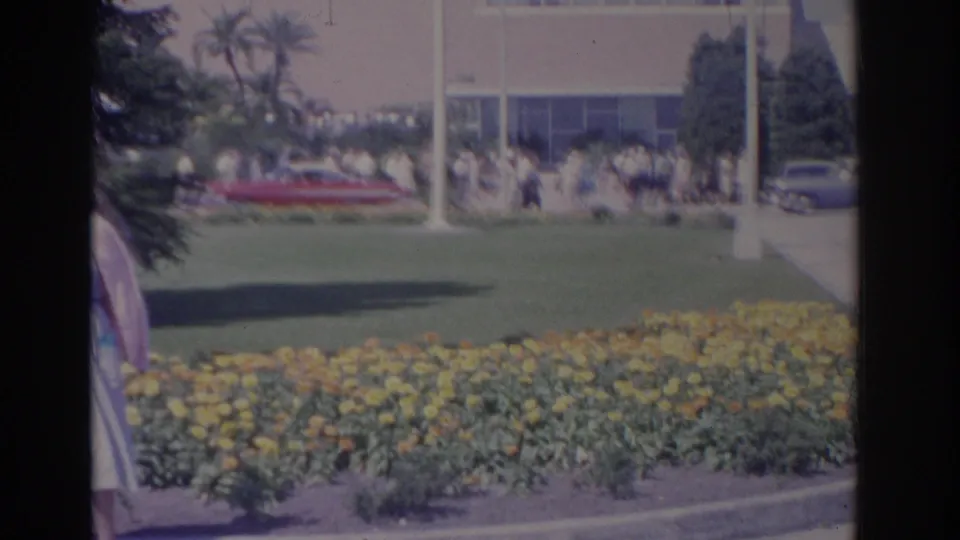 The camera drifts so the woman is mostly cropped at the edge, emphasizing the yellow flowers, open lawn, building, visitors, and parked cars.
