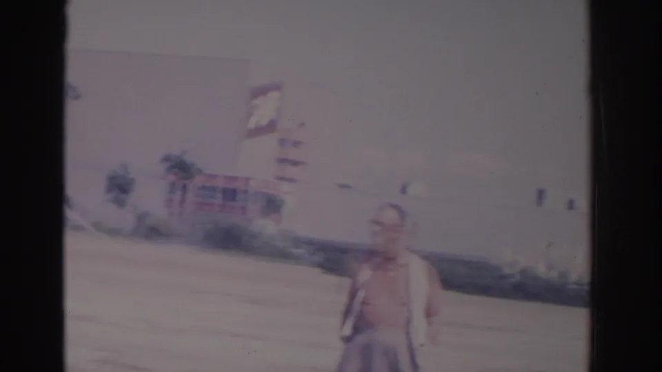 1962 Florida frame of a man near the roadside, with a large building and a partly visible Schlitz sign in the background.