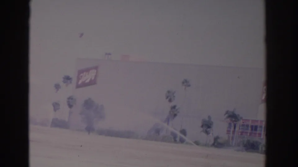 1962 Florida 8mm frame showing a large building with a Schlitz sign, palm landscaping, sprinkler spray, and sunlit pavement.